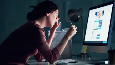 A woman sits in front of a large computer screen and rubs at her eyes