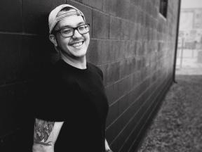 man with glasses and hat smiling in grayscale leaning against brick wall