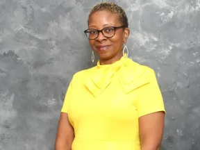 Photo of woman in yellow standing in front of gray background.