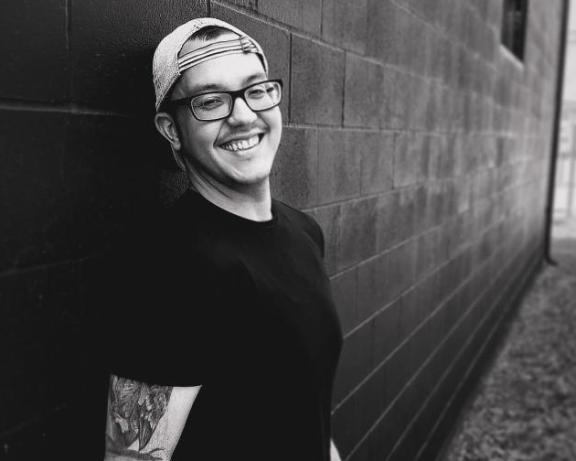 man with glasses and hat smiling in grayscale leaning against brick wall