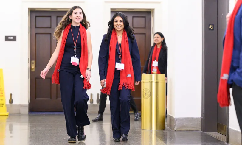 Two smiling women in red scarves and ID badges walk confidently in a hallway.