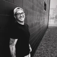 man with glasses and hat smiling in grayscale leaning against brick wall