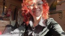 A selfie of a person with curly red hair and glasses smiling into the camera.