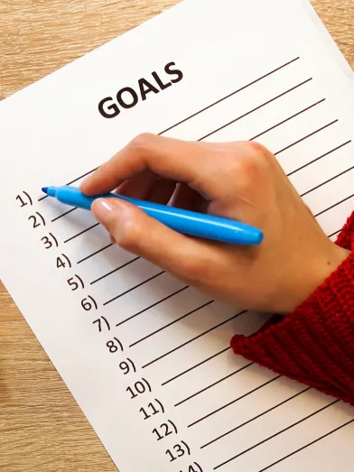 A hand holding a blue pen prepares to fill out a list of goals. 