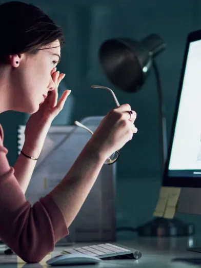 A woman sits in front of a large computer screen and rubs at her eyes